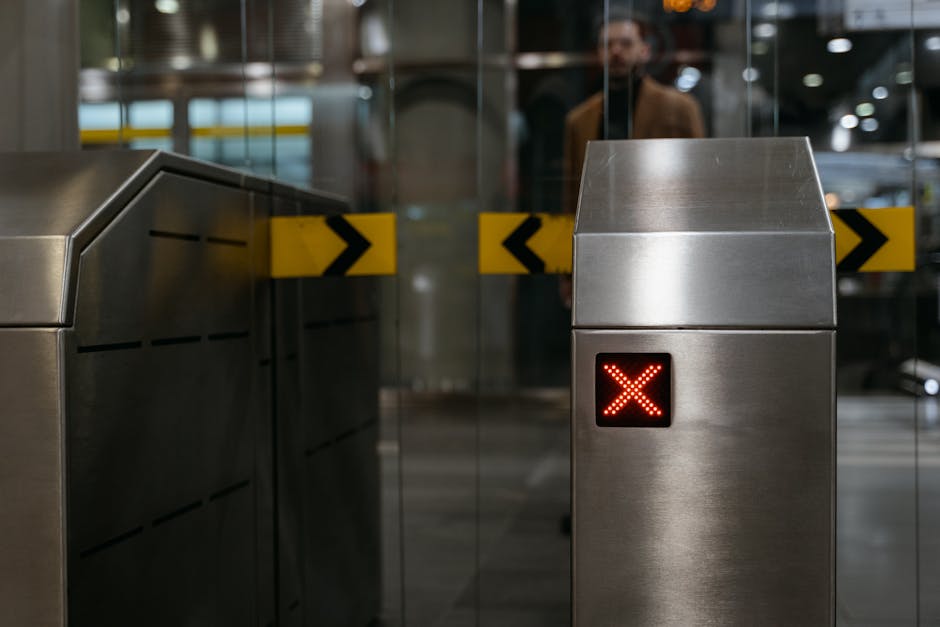 Image of an indoor airport or train station entrance featuring a metal access control barrier with a red cross digital indicator, indicating restricted access. Behind the barrier, a glass sliding door is visible, with a blurred figure of a person walking in the background. The scene is well-lit with artificial lighting, and there are yellow and black safety tape markings on the barrier, likely indicating a controlled or restricted zone. The metal barrier and the digital display suggest a secure environment, possibly related to travel or transportation. This setting may relate to the logistical aspects of home relocation or furniture transport, as part of a professional removals service like Removal Van Kensington, operating within complex transit environments for house moving processes.
