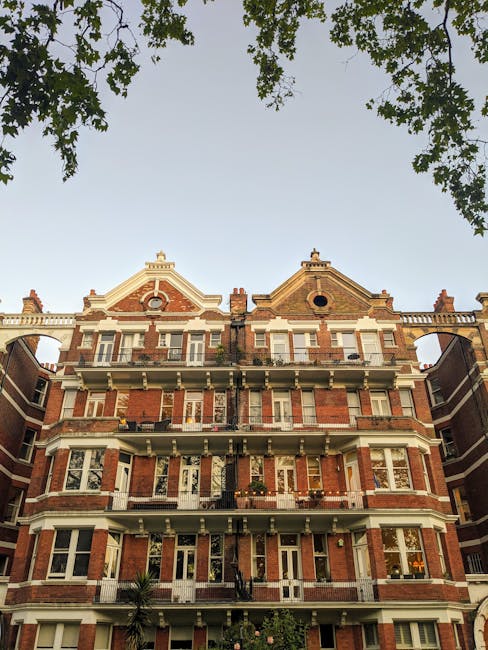 An exterior view of a historic red-brick apartment building with ornate architectural details, featuring four floors and multiple windows with white trims. The ground floor shows a doorway with black steps leading inside, and a small potted plant is visible near the entrance. The building has balconies with black iron railings on each floor, some with visible furniture or decorative items. The roofline includes decorative gables and chimneys, and the sky above is clear with a warm light, suggesting daytime. In the foreground, the tops of green trees frame the upper part of the image, partially obscuring the sky. As part of a house removal or relocation project, the premises are clean and well-maintained, illustrating the type of property typically serviced by Removal Van Kensington during furniture transport and home relocation tasks in Holland Park, Kensington.
