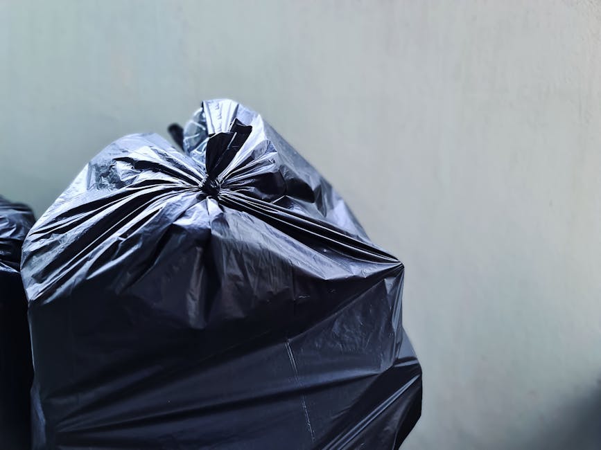 A person wearing a white T-shirt and transparent disposable gloves is holding a white plastic bin labeled 'PLASTIC' in black marker. The bin contains a black rubbish bag lined along the top, with a tr