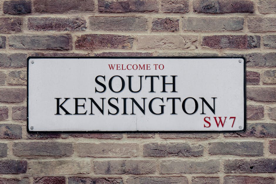 Close-up of a rectangular white street sign mounted on a red brick wall, displaying the text 'Welcome to South Kensington' with 'SW7' in the bottom right corner in red. The sign features black and red lettering and is secured with visible screws. The brick wall behind the sign shows a pattern of horizontal bricks with various shades of red and brown, indicating an outdoor setting typical of residential or commercial properties in London. This signage can be seen as part of the context for a house removal or home relocation service, such as those provided by Removal Van Kensington, highlighting the area where furniture transport and packing and moving operations might take place during a house move in South Kensington.