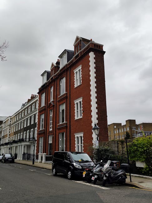 A multi-story brick building on a city street with a decorative white trim running vertically along the corner and window frames. The building features large sash windows, some with shutters, and a gabled roof with dormer windows at the top. Adjacent buildings of varying heights and architectural styles line the street, which is paved and has parked vehicles, including a black van and several motor scooters, close to the curb. The scene is set under a cloudy sky, with the environment suggesting a residential area in Kensington, suitable for house removals and home relocation services by Removal Van Kensington as part of their furniture transport and packing and moving operations.