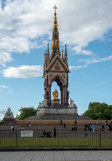 A tall, ornate monument with intricate Gothic-style details and a golden figure at its base, situated outdoors in a park-like setting with a partly cloudy sky overhead. The structure features a spire with decorative elements, arches, and statues, and is surrounded by a small fence. In the foreground, there are several people, some walking or sitting on benches, and others gathered near the base of the monument. The scene captures a clear day with natural light highlighting the architectural details. The environment suggests a popular public space, possibly in central London, where Removal Van Kensington provides house removal and furniture transport services, often involving the careful packing and loading of household items for relocation.