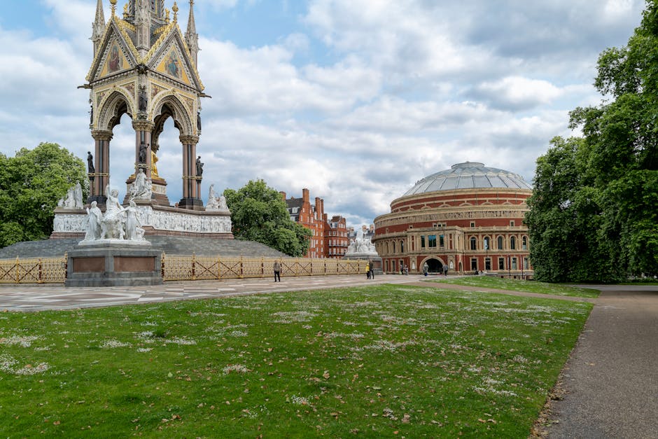A view of a bright day in a park with a statue and monument featuring intricate Gothic architectural details, positioned on a paved area with decorative gold fencing. To the right, there is a large, round, red-brick building with a domed roof and arched windows, surrounded by lush green trees. In the foreground, a grassy area with scattered white flowers and a paved pathway extends towards these structures. The scene suggests an outdoor setting linked to cultural or historic landmarks within a London park, aligned with the context of house removals and furniture transport by Removal Van Kensington.