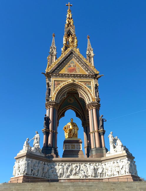 A tall, ornate monument with intricate Gothic-style details and a golden figure at its base, situated outdoors in a park-like setting with a partly cloudy sky overhead. The structure features a spire with decorative elements, arches, and statues, and is surrounded by a small fence. In the foreground, there are several people, some walking or sitting on benches, and others gathered near the base of the monument. The scene captures a clear day with natural light highlighting the architectural details. The environment suggests a popular public space, possibly in central London, where Removal Van Kensington provides house removal and furniture transport services, often involving the careful packing and loading of household items for relocation.
