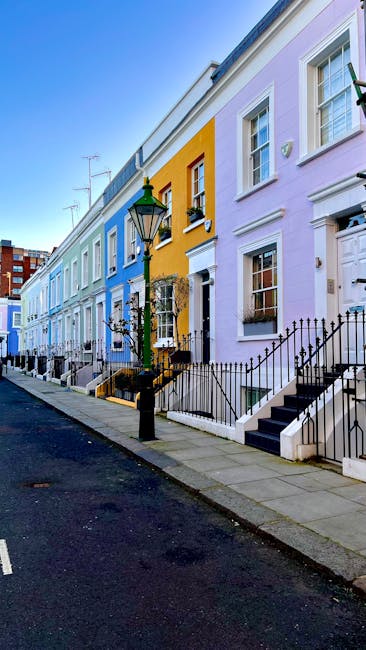 A row of colorful terraced houses in a residential street during daytime, with pastel facades painted in shades of blue, yellow, and light purple. The houses feature white-framed windows, some with flower boxes, and small front steps leading up to white doorways. A traditional black metal fence runs along the sidewalk in front of the houses, with a street lamp positioned near the pavement. The street is empty, with a parked van partially visible in the background. Inside the property, a removal van from Removal Van Kensington is parked nearby, with an open doorway showing a cluttered interior containing cardboard boxes, bubble wrap, and plastic wrapping materials. Furniture such as a wooden chair, a small table, and packed boxes wrapped in plastic and fabric are being loaded into the van as part of a home relocation or furniture transport process. The lighting indicates clear weather, and the scene captures a typical urban setup involved in packing, loading, and planning a house move, illustrating the logistical elements of same-day removals in Kensington.