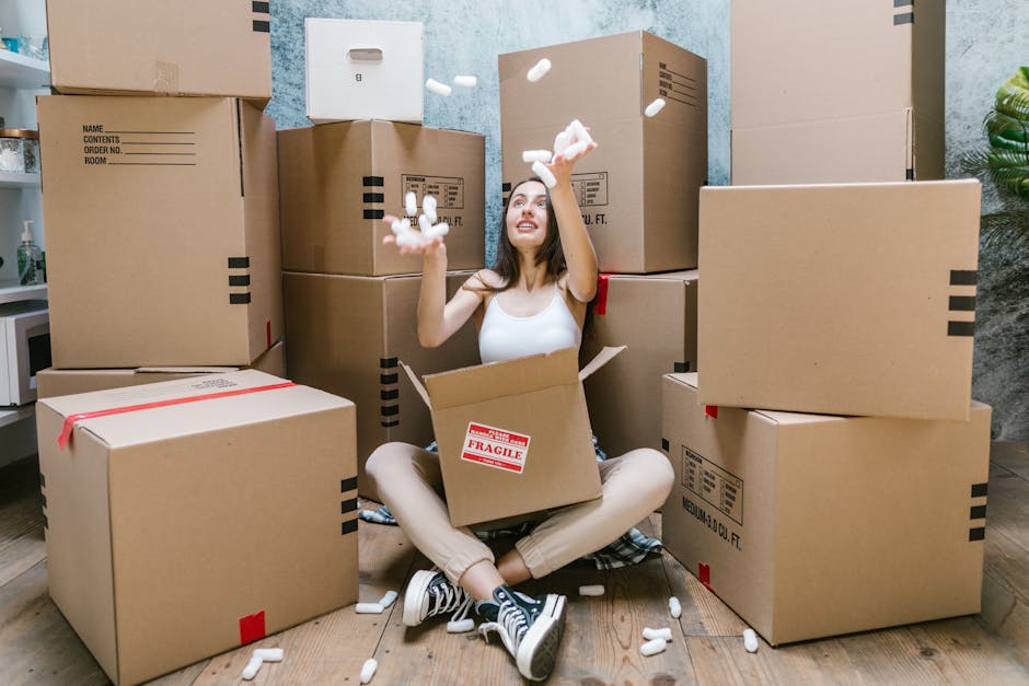 A young woman with long brown hair, sitting on a wooden floor inside a room, surrounded by numerous large cardboard moving boxes stacked around her. She is inside one open box, dressed casually in a white sleeveless top, beige pants, and black-and-white sneakers. She is smiling and tossing packing foam peanuts into the air, with some falling around her. The boxes are taped and some have labels, indicating they are packed for a home relocation. The room has light-colored walls and a potted plant visible on the right side, with natural lighting illuminating the scene. This image depicts packing and moving preparations, related to furniture transport and loading in the context of house removals, which aligns with services offered by Removal Van Kensington.