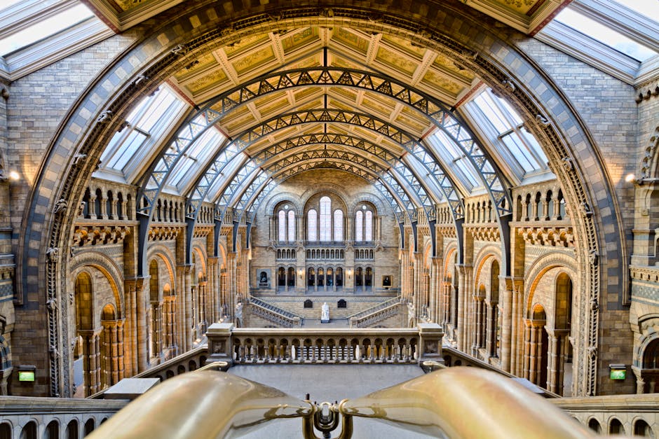 The image depicts the interior of a historic, grand hall with a high, arched ceiling supported by ornate metal framework and large, arched windows allowing natural light to illuminate the space. The walls are built with stone bricks and feature decorative arches and columns. A wide staircase with stone balustrades leads down to the central level where a statue is visible in front of a series of tall, narrow windows. The spacious area appears suitable for a museum or gallery, with no furniture or objects related to house removals visible. This setting is associated with historic architecture, possibly related to the context of house or museum relocations, as referenced by the title 'South Kensington removals for museum moves, KENSINGTON.' The overall scene emphasizes the structural details and grandeur of the building, implying careful handling during transport or moving services.